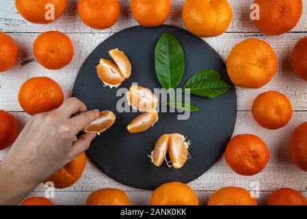 Human hand is taking tangerine segments from round black plate surrounded by whole orange tangerines on old white wooden table. Fruits have natural im Stock Photo