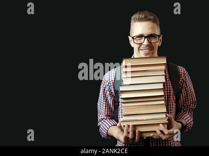 Front view of smiling student with glasses and backpack carrying many textbooks isolated on dark background Stock Photo