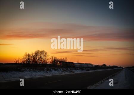 An industrial freight train illuminated by headlights travelling at sunset along a deserted highway in a prairie winter landscape Stock Photo