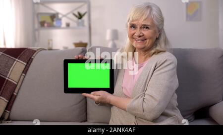 Smiling mature lady holding green screen tablet in hands, easy banking, shopping Stock Photo