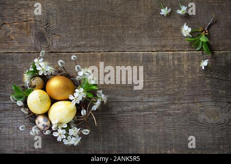 Golden Colored easter eggs in a nest with willow branches and spring flowers on a gray wooden background. Top view flat lay background. Copy space. Stock Photo