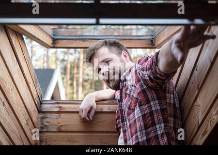 Handsome bearded man stands near an open skylight in the attic. Stock Photo