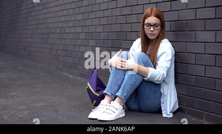 Lonely female student eyeglasses sitting floor in high school backyard, problem Stock Photo