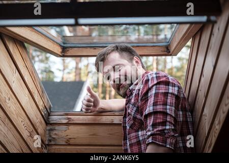 Bearded man stands near an open roof window in attic and makes an approval gesture, thumb up, smiling and looking at the camera. Stock Photo