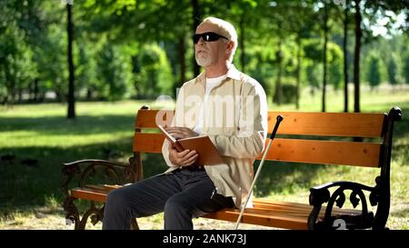 Blind pensioner reading braille book, sitting on bench in summer park, resting Stock Photo