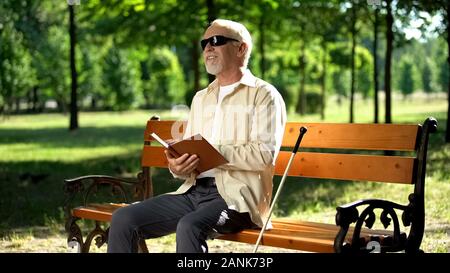Happy blind pensioner reading funny story in braille book, enjoying rest in park Stock Photo