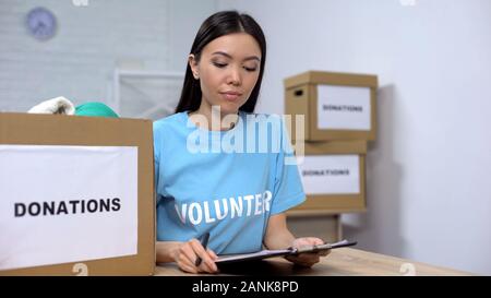 Young volunteer checking clothes donation box and making notes, humanitarian aid Stock Photo