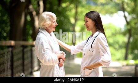 Smiling nurse talking with elderly woman in bathrobe at sanatoria park, relax Stock Photo