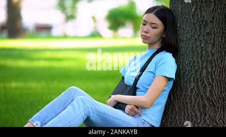 Sad lady with injured arm in supporting sling sitting tree in park, medicine Stock Photo