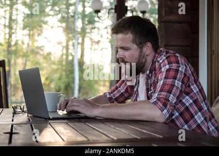 Bearded man works with a laptop on the outdoor terrace in his yard, against the background of trees. Stock Photo