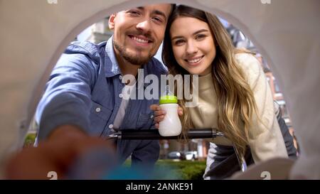 Smiling parents putting baby pacifier to carriage, mum holding milk bottle Stock Photo