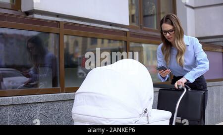 Multitasking business mother answering message as walking to work with baby pram Stock Photo