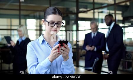 Pretty businesslady looking at smartphone, colleagues in office, business app Stock Photo