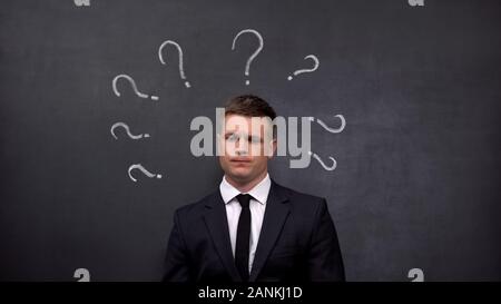 Doubtful businessman lacking ideas, question marks painted on blackboard, answer Stock Photo