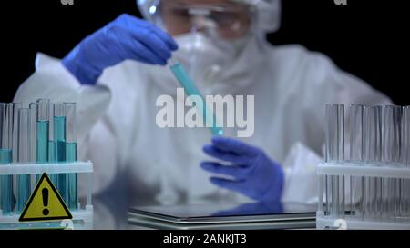 Scientist in protective suit analyzing dangerous liquid tubes, tablet on table Stock Photo