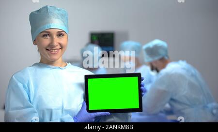 Smiling nurse holding tablet PC with green screen during operation, hospital ad Stock Photo
