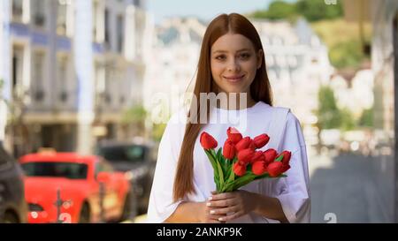 Happy teenage red-haired girl holding tulips bouquet and smiling on camera, gift Stock Photo
