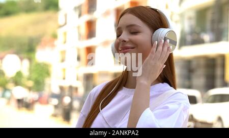 Pretty female teenager in headphones listening to favorite music on city street Stock Photo