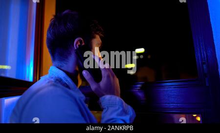 Sad man talking on phone and looking through window, waiting for wife from work Stock Photo