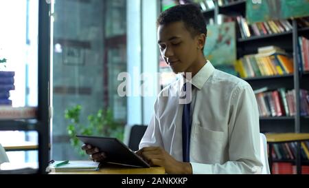 Geek mixed-race male student doing homework online, using tablet pc at library Stock Photo