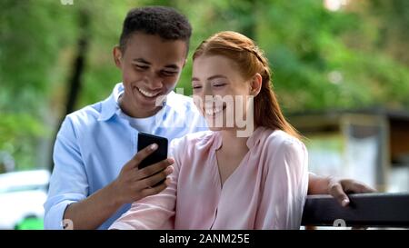 Smiling couple of teenagers watching photos on smartphone sitting on bench Stock Photo