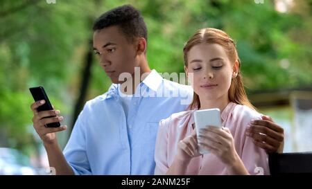 Hugging multiethnic teen couple sitting on bench in park, chatting smartphone Stock Photo
