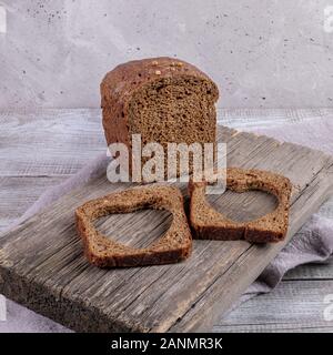 Loaf of rye bread and two slices with carved holes of heart shape in them on old wooden Board on grey background. Healthy leavened bread. Valentine's Stock Photo