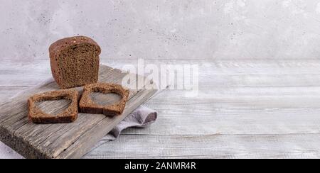 Loaf of rye bread and two slices with carved holes of heart shape in them on old wooden Board on grey background. Valentine's or mother's day concept. Stock Photo