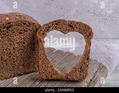 Loaf of rye bread and slice with carved hole of heart shape in it on old wooden board on grey concrete backdrop. Healthy leavened bread. Valentine's o Stock Photo