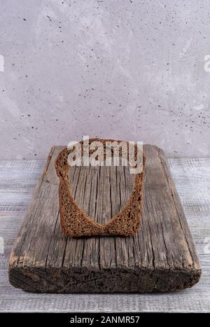 One rye bread slice with carved hole of heart shape in it on old wooden board on grey concrete backdrop. Healthy leavened bread. Valentine's or mother Stock Photo