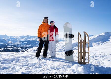 Portrait Of A Happy Young Couple Standing Near Snow Board And Wooden Sledge In Snow Stock Photo