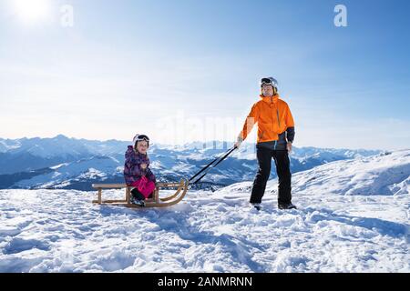 Young Man Looking At Camera While Pulling His Daughter On Wooden Sledge In Snow Stock Photo
