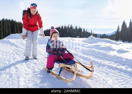 Adorable Girl Enjoying Sledding While Her Mother Pushing Sledge Against Pine Tree Forest During Winter Stock Photo