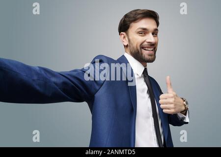 wide angle selfie shot of young business man going thumb up on grey background Stock Photo