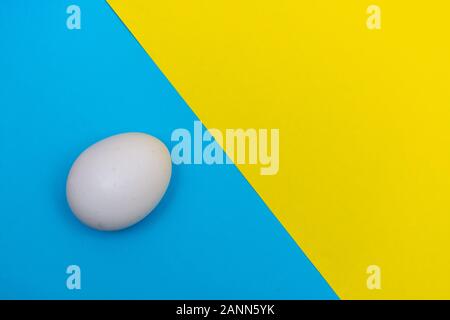 Easter holiday flat lay with white egg on a solid bright blue and yellow vibrant background with copy space. Stock Photo