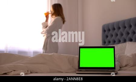 Laptop on bed, woman drinking juice relaxing near window, online travel booking Stock Photo