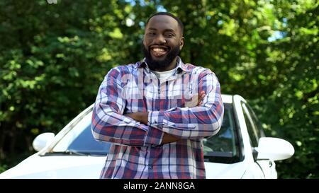 Happy afro-american man smiling on cam, leaning new automobile, rent service Stock Photo