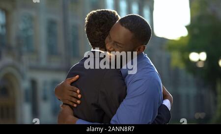 Cheerful black father embracing young son in prom suit, college graduation, joy Stock Photo