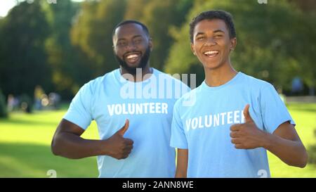 Two joyful american men in volunteer t-shirts showing thumbs up, selfless help Stock Photo