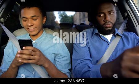 Glad teenager chatting phone in car, dad trying to read messages, parent control Stock Photo