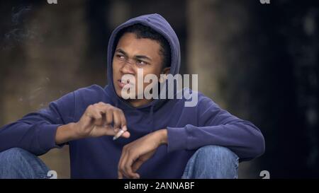 Difficult teenage boy smoking cigarette sitting street, lack of parental control Stock Photo