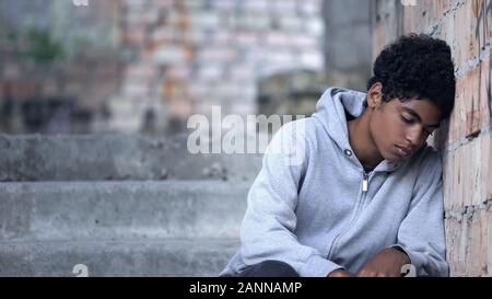 Depressed male teenager leaning brick wall sitting stairs, domestic violence Stock Photo