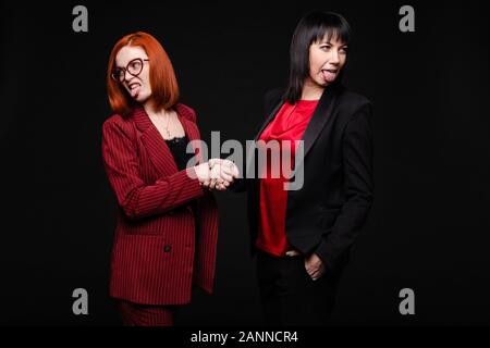 Stock photo of two businesswomen in red and black suits shaking hands and looking in different directions and sticking tongues out. Business partners Stock Photo