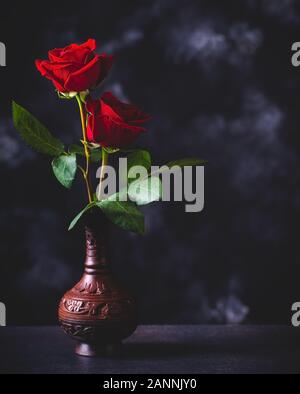Two red roses in a vase on a dark surface and background Stock Photo