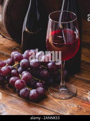 Glass of red wine and grapes with two wine bottles and wooden barrel in background Stock Photo