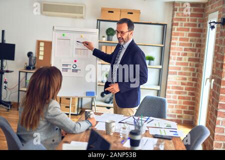 Two middle age business workers smiling happy and confidentt working together with smile on face. Man standing doing work presentation using board and Stock Photo