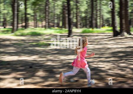 A blond girl wearing a pink outfit runs through the forest on a sunny day. Her curly hair bounces with the wind of her speed. Stock Photo