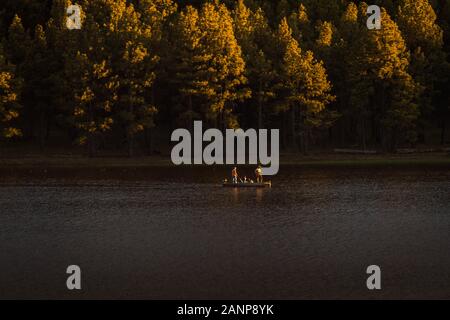 Two men and their kids are on a boat fishing at Lake Mary in Flagstaff, Arizona. The pine forest behind them is highlighted with gold sunlight. Stock Photo