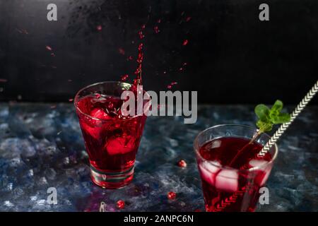 Pomegranate juice in transparent glasses. Spray from a falling piece of ice. On a blue background under a stone. Trend 2020. Grains are scattered near Stock Photo