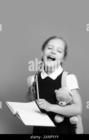 Kid in school uniform isolated on green background. Study and back to school concept. Pupil holds blue book, marker and teddy bear. Girl with braids with open mouth and closed eyes Stock Photo